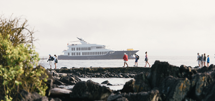 Tourists walk on the lava rock coastline of the Galápagos Islands, with a luxury yacht in the background