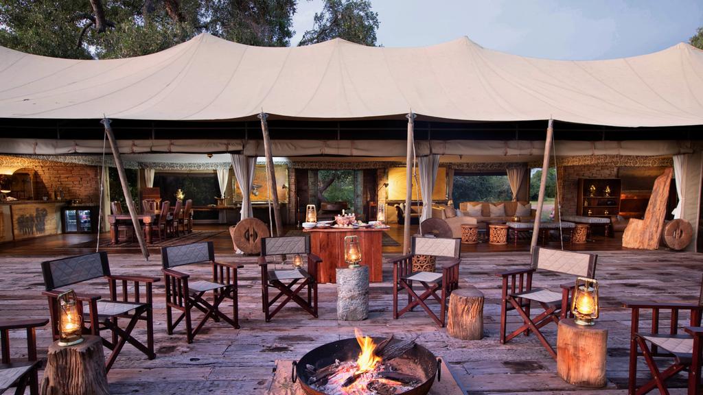 Outdoor lounge area at Tembo Plains Camp in Sapi Reserve, Zimbabwe, featuring a campfire surrounded by canvas safari chairs on a wooden deck with an open dining and bar area in the background.
