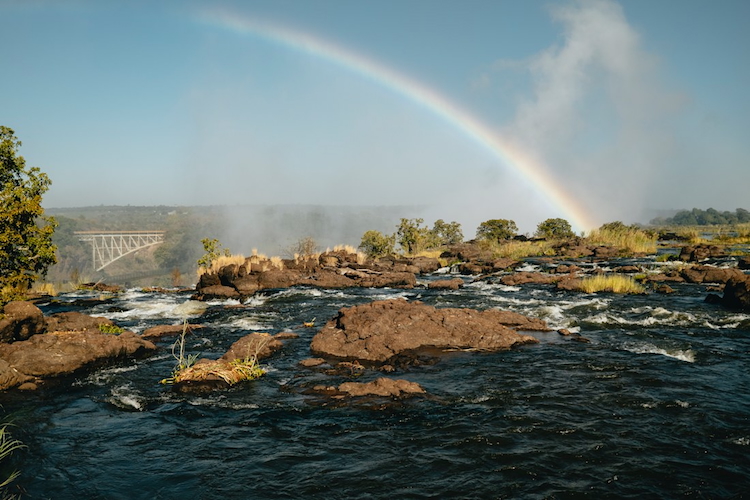 Rainbow over Victoria Falls with spray mist and Zambezi River rapids in the foreground, and the Victoria Falls Bridge in the distance.