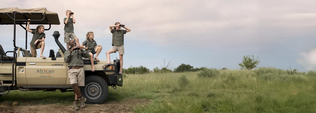 Excited children on safari climb on a game vehicle with their guide at Somalisa Acacia, scanning the plains with binoculars.