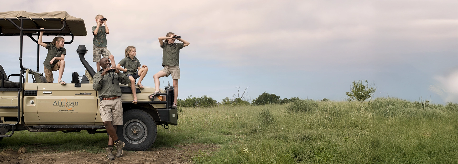 Excited children on safari climb on a game vehicle with their guide at Somalisa Acacia, scanning the plains with binoculars.