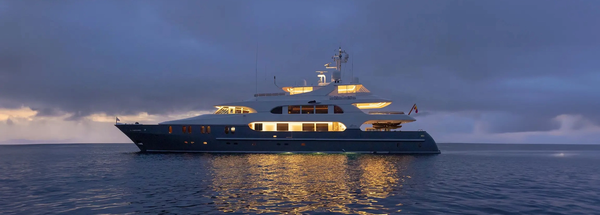 Aqua Lares yacht illuminated at dusk, its golden lights reflecting softly across calm Indian Ocean waters beneath a brooding sky.