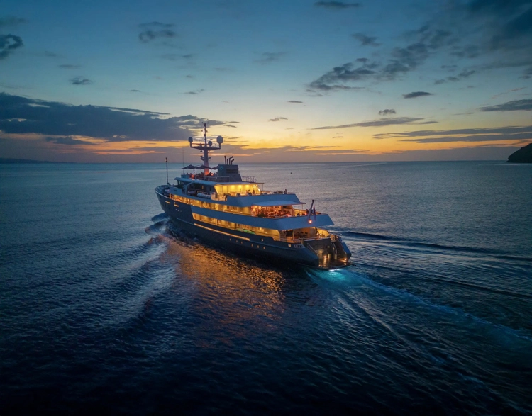 Aqua Blu yacht illuminated at twilight as it sails through calm Indonesian waters beneath a fading sunset.