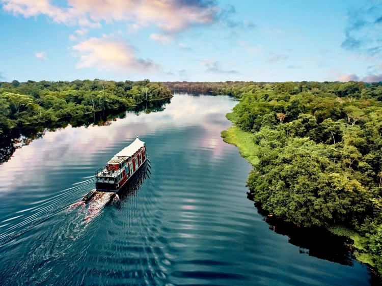 Aria Amazon river vessel gliding along a tranquil jungle waterway framed by lush rainforest and pink clouds.