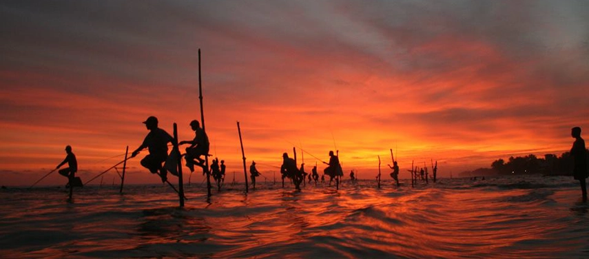 Traditional Sri Lankan stilt fishermen silhouetted against a dramatic red-orange sunset sky.