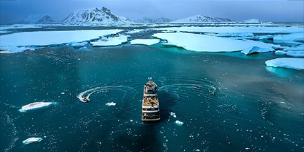 Aerial view of Aqua Lares sailing between drifting ice floes under soft Arctic light.