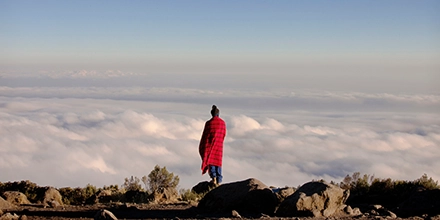 Maasai man in traditional red shuka standing above a sea of clouds on Mount Kilimanjaro.