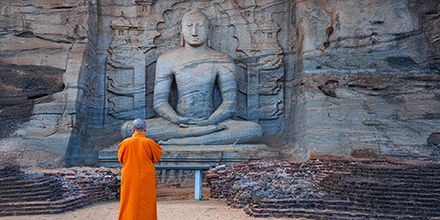 Buddhist monk in orange robes standing before a giant rock-carved seated Buddha statue in Sri Lanka.