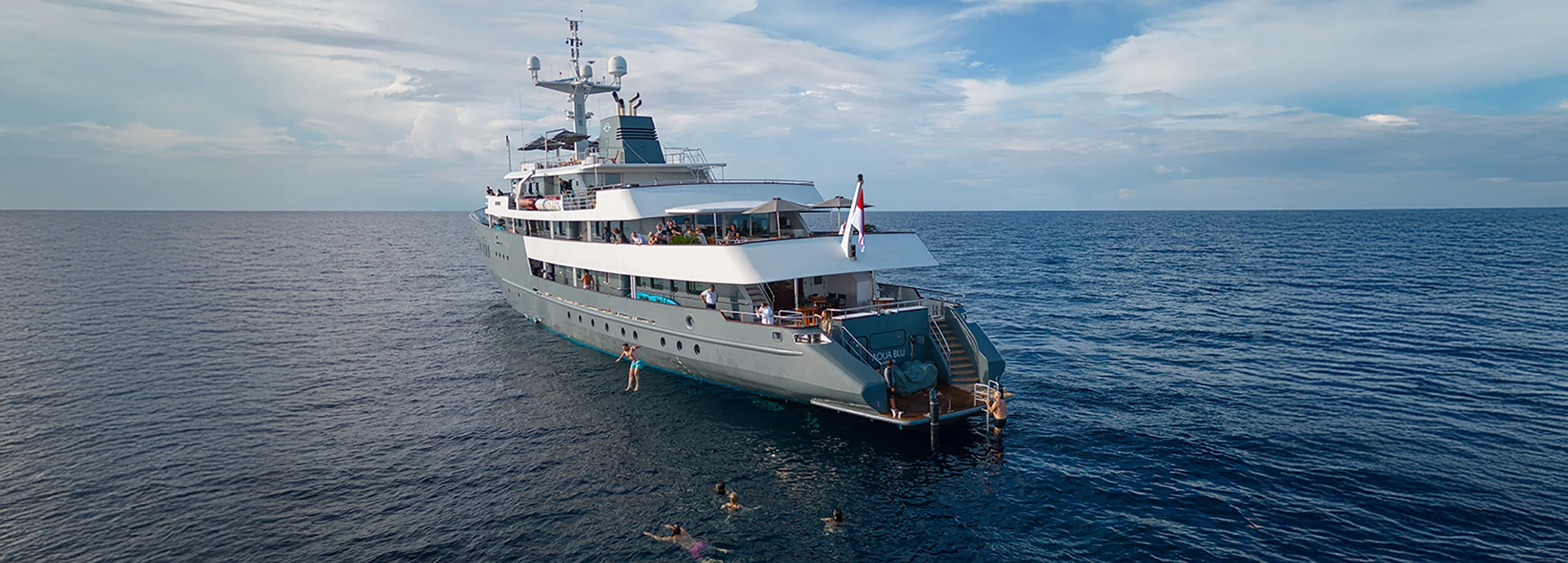 Travellers swim beside Aqua Blu in the open sea, leaping from the yacht’s deck into clear tropical waters.
