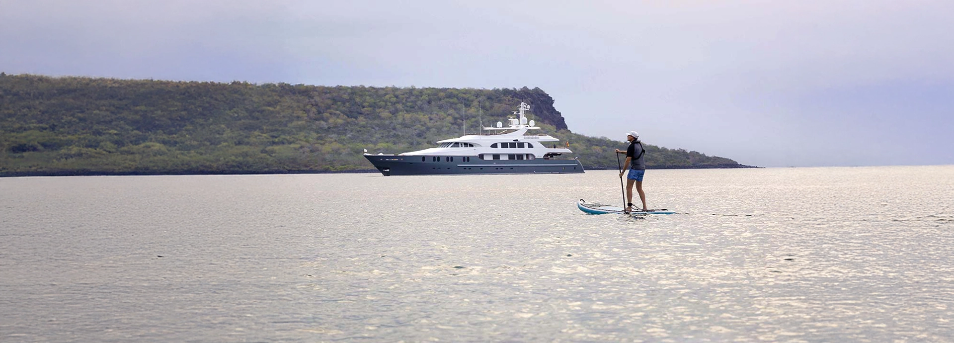 A paddleboarder glides across tranquil Galápagos waters, with Aqua Mare anchored nearby beneath pale island skies.