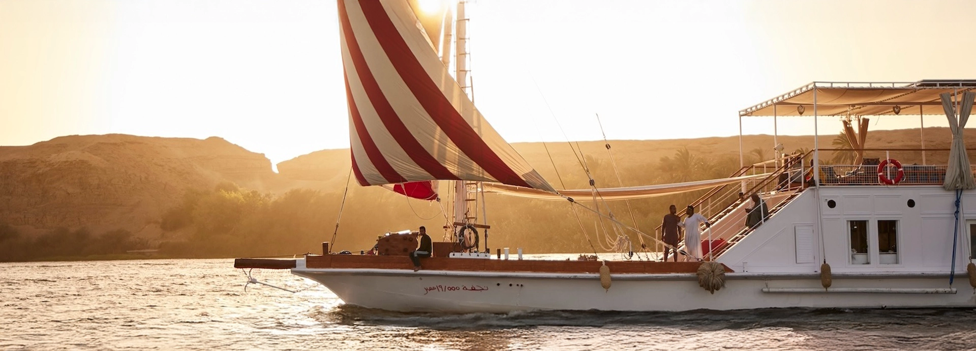A dahabiya sweeps along the river at dusk, its sails glowing as the sun sets over the desert hills.