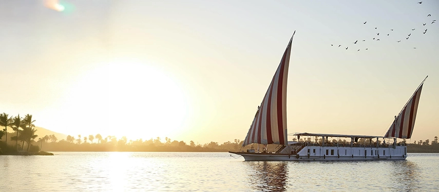 A dahabiya glides through shimmering morning water, its sails lit by the rising sun over the river’s edge.