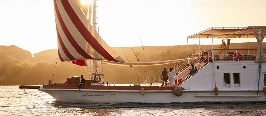 A traditional dahabiya sails through glowing sunset light, its elegant lines mirrored on the Nile’s rippling surface.
