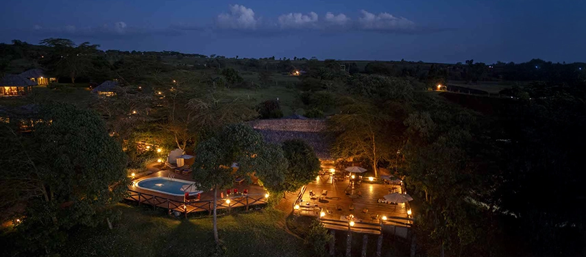 Twilight aerial view of Karatu Simba Lodge in Tanzania, with warm lighting around the pool and outdoor dining area nestled in forested surroundings.