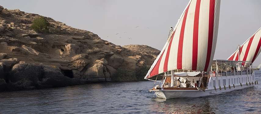 A dahabiya slips past rocky cliffs, its striped sails bright against the warm desert stone.