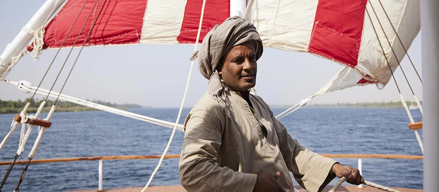 A crewman guides the dahabiya across the Nile, the striped sails billowing softly in the warm morning breeze.