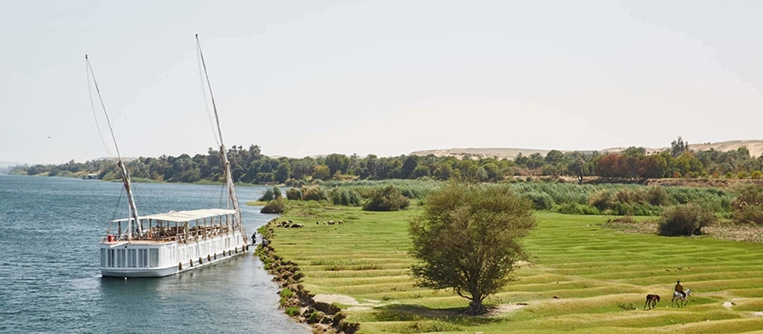A dahabiya rests beside lush riverside fields where farmers and animals move through a peaceful green landscape.