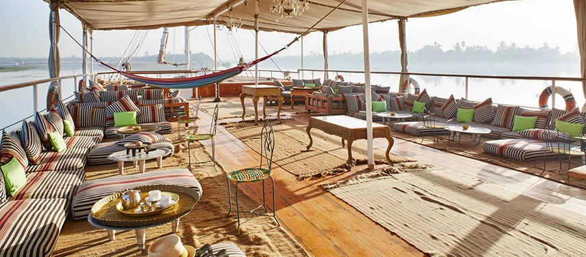 The broad sun deck stretches beneath a billowing canopy, its striped cushions and hammocks inviting long hours of river-side relaxation.