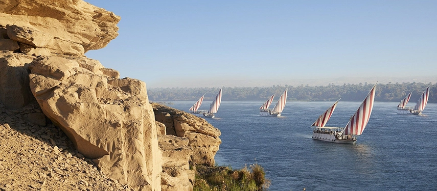 A fleet of dahabiyas move gracefully along the Nile, framed by rocky cliffs and a soft morning haze.