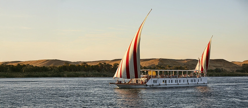 A dahabiya glides along the Nile at golden hour, its striped sails catching the warm desert breeze.