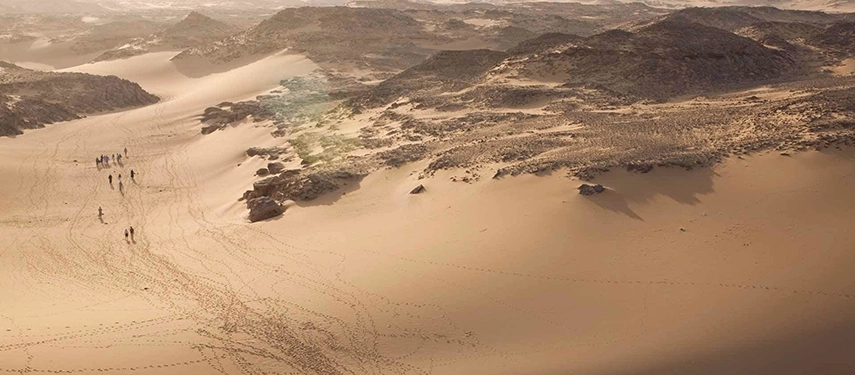 Travellers walk across soft desert dunes under warm afternoon light, surrounded by vast, sculpted landscapes.