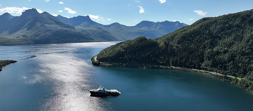 Aqua Lares cruising through a deep fjord framed by emerald hills and distant mountains.