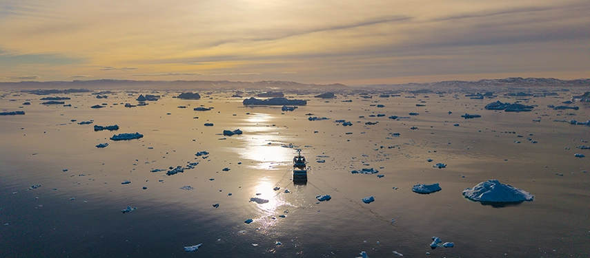 Aqua Lares sailing beneath a glowing Arctic sunset across a sea scattered with icebergs.