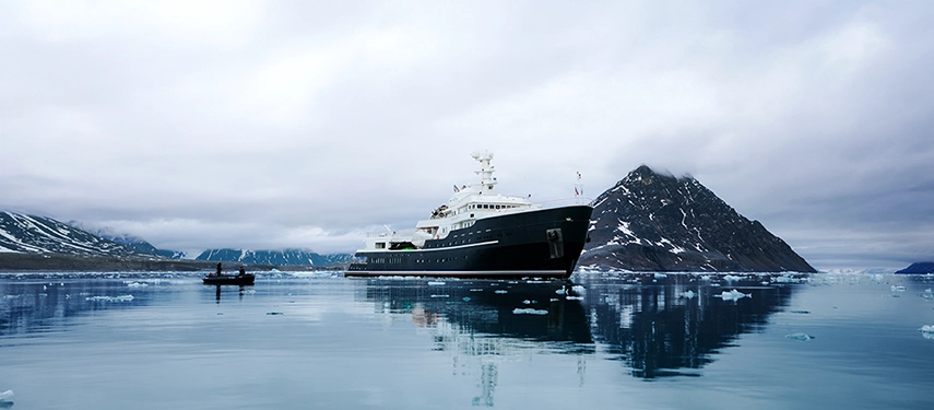 Aqua Lares anchored near a glacier-lined shoreline under a soft grey Arctic sky.