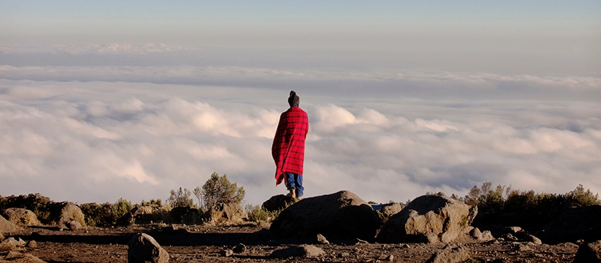 Maasai man in traditional red shuka standing above a sea of clouds on Mount Kilimanjaro.