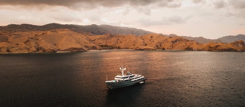 Aqua Blu anchored in a calm bay framed by sunlit Indonesian hills, her reflection mirrored in the gentle sea.