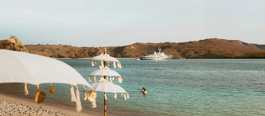 White parasols line a tranquil beach as Aqua Blu floats offshore, the perfect setting for an elegant island sundowner.