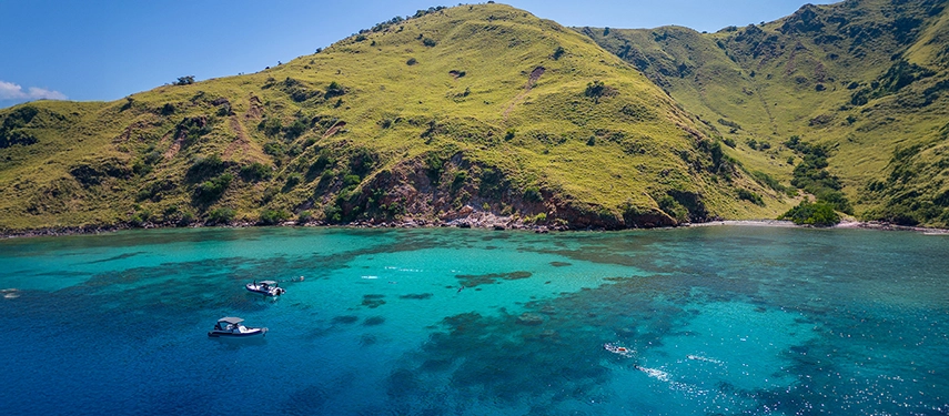 Snorkellers float above glowing coral gardens in shallow turquoise water near a remote Indonesian island.