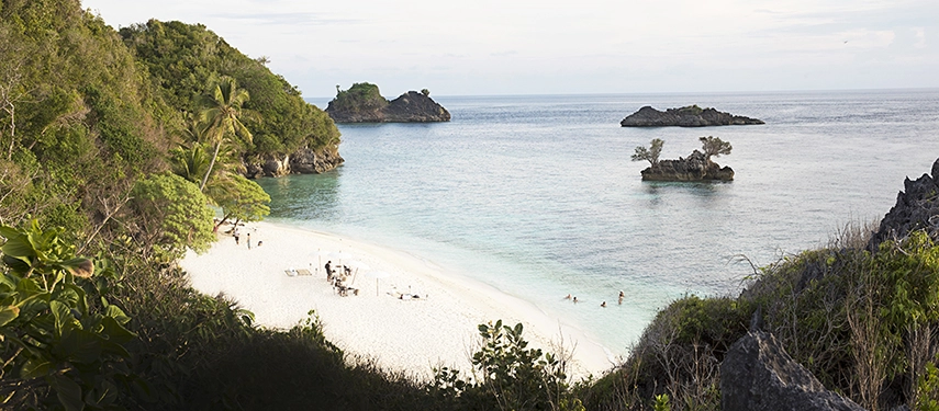 Secluded white-sand beach fringed by lush jungle and turquoise water, with small rock islets rising just offshore.