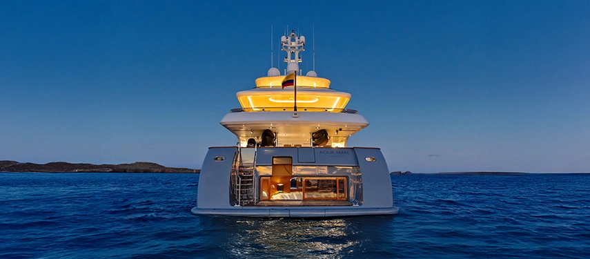 The illuminated stern of Aqua Mare glows against the evening sea, revealing the beach club’s water-level suite and sleek super-yacht design.