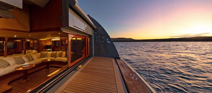 The beach club’s teak deck extends to the ocean, with soft lighting and open glass doors revealing the yacht’s elegant interior salon.