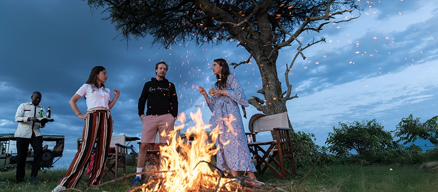 Guests gathered around a glowing campfire under a tree at dusk at Basecamp Masai Mara, enjoying drinks and conversation as embers float into the air.
