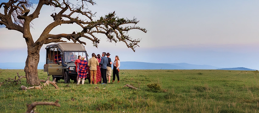 Group of guests and Maasai guides enjoying a bush stop beside a safari vehicle under a lone tree, with vast Masai Mara plains stretching to the horizon.