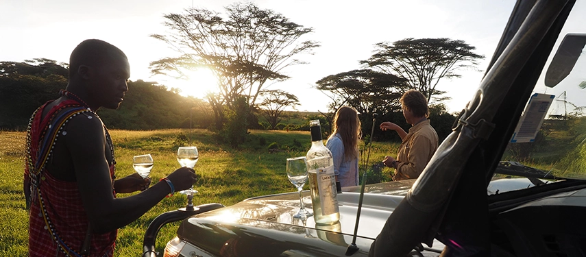 Maasai guide serving sundowners to guests beside a safari vehicle, as golden sunlight filters through acacia trees at Basecamp Masai Mara.