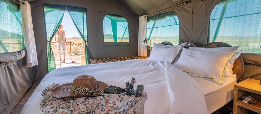 Interior of a luxury safari tent at Camp Doros featuring a neatly made bed, rustic wooden furniture, and scenic views through screened windows.