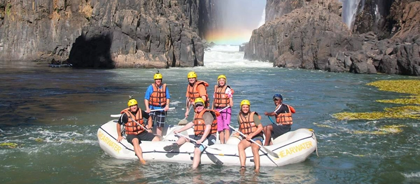 Group of adventurers posing in a whitewater rafting boat with a rainbow forming near the base of Victoria Falls on the Grand Zambezi Safari.