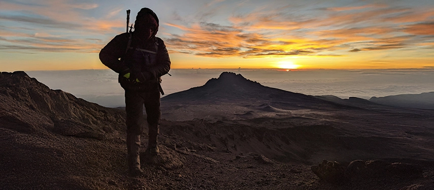 Silhouetted trekker walking across volcanic slopes with Mount Mawenzi and a vivid sunrise lighting the horizon.