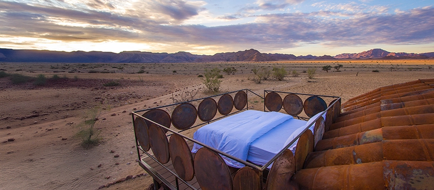 Outdoor sleepout deck at Camp Sossus featuring a raised bed surrounded by repurposed metal drums overlooking the Namibian wilderness.