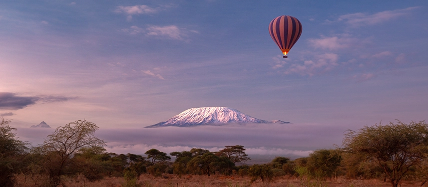 Hot air balloon floating at dawn above the savannah with Mount Kilimanjaro rising above a low sea of clouds.