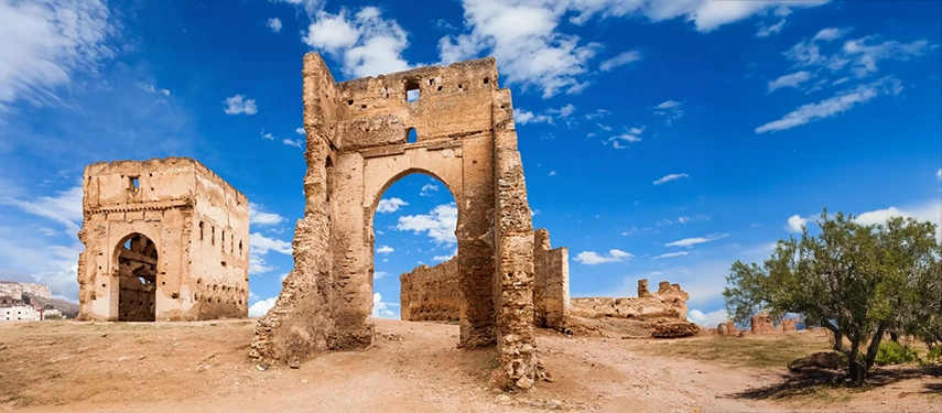 Ancient ruins of the Merenid Tombs overlooking Fès, with weathered stone arches beneath a bright blue sky.