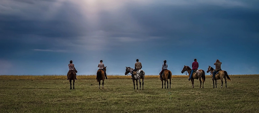 Six riders on horseback silhouetted against a dramatic Zambian sky over wide open grasslands.