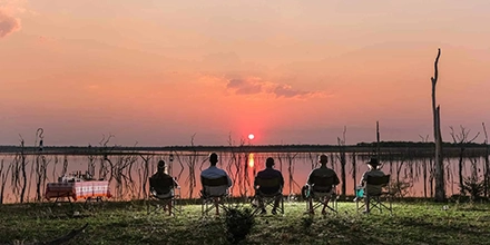 Guests seated at sunset facing Lake Kariba, watching the sun dip behind flooded trees rising from the water.
