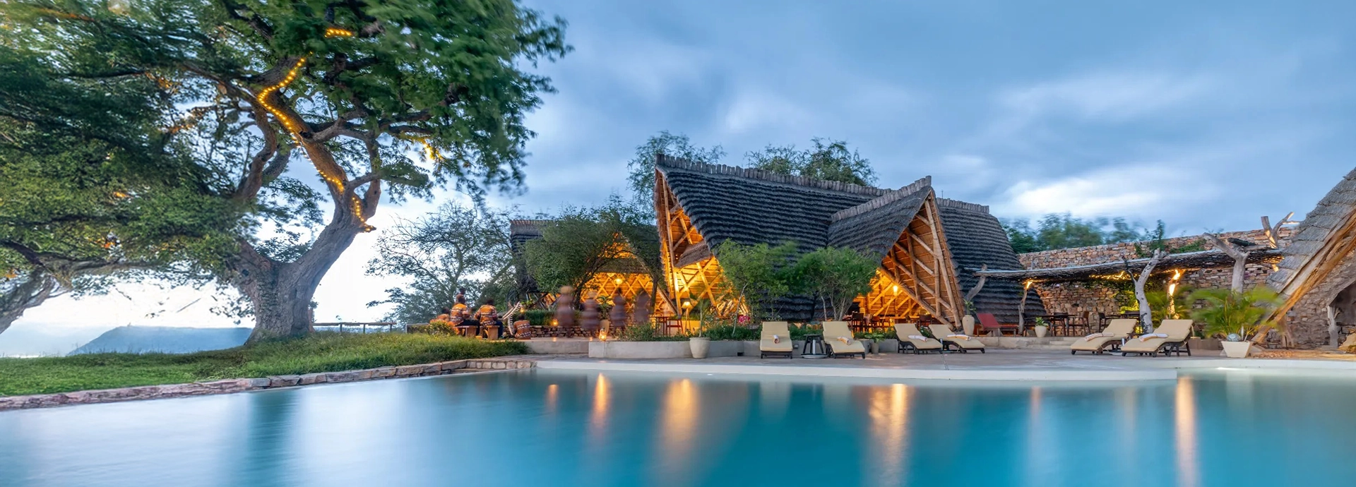 The lodge swimming pool reflects the evening sky, with thatched buildings and softly lit communal spaces creating a tranquil riverfront atmosphere.